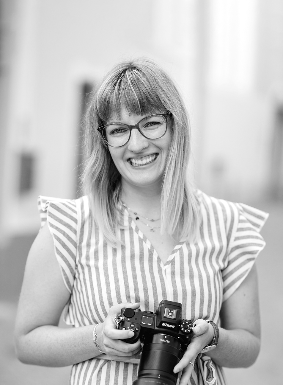 Das Bild in schwarz weiß zeigt eine junge grinsende Frau, die eine Kamera nach unten gerichtet in der Hand hält. Sie trägt eine Brille, einen Pony und hat offene lange Haare, die ihr bis zu den Schlüsselbeinen reichen. Sie trägt ein gestreiftes Oberteil mit kurzen rüschenartigen Ärmeln an den Schultern.