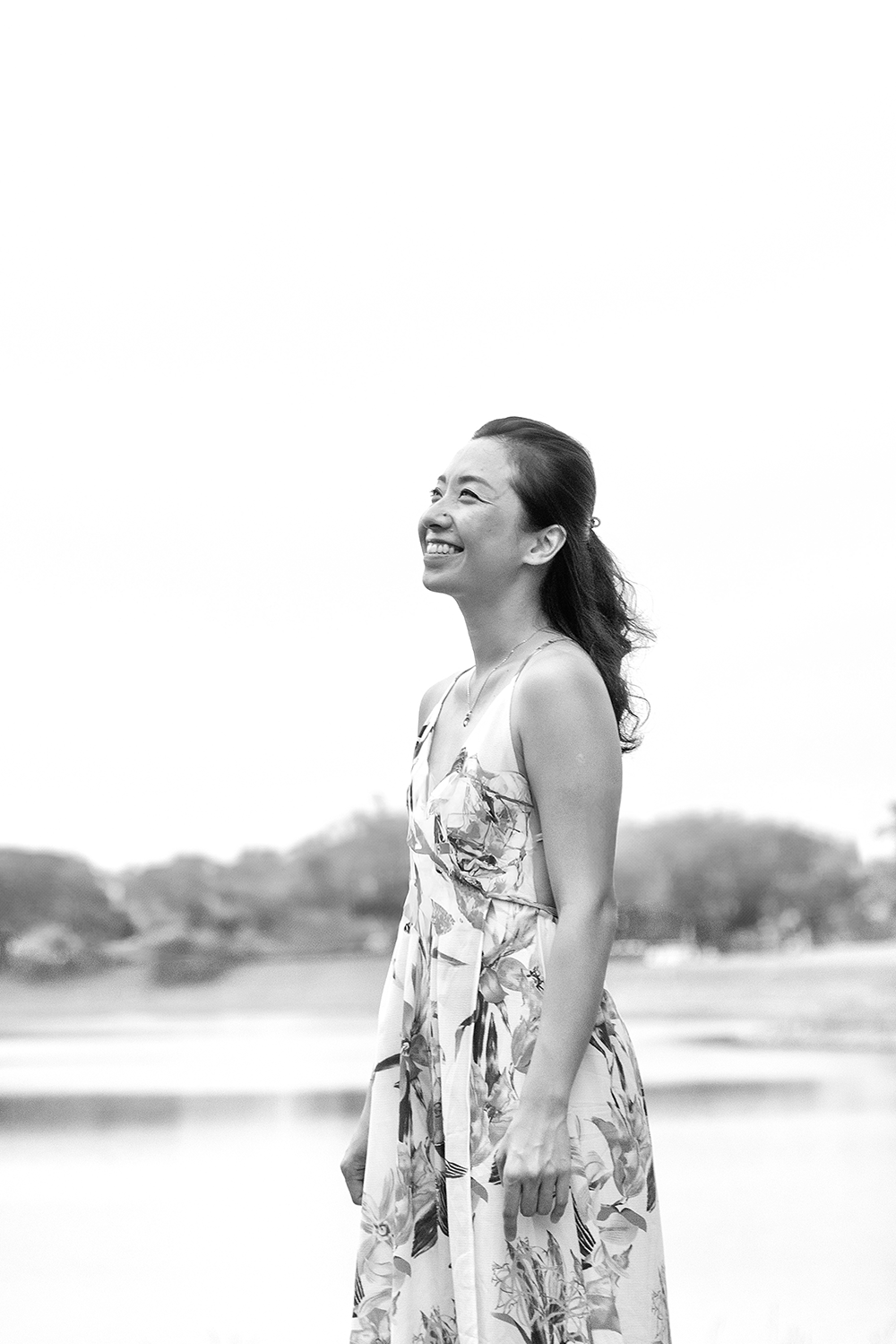 Das schwarz weiß Bild zeigt eine junge Frau in der Natur, die mit einem freudigen Gesichtsausdruck in die Luft schaut. Sie hat einen asiatisch kulturellen Background und trägt ihre langen schwarzen Haare nach hinten gebunden, sodass sie ihr offen über den Rücken fallen. Sie trägt ein Ärmelloses langes und schickes Kleid, hinter ihr in der Ferne sieht man Hügel, Wald und einen See.