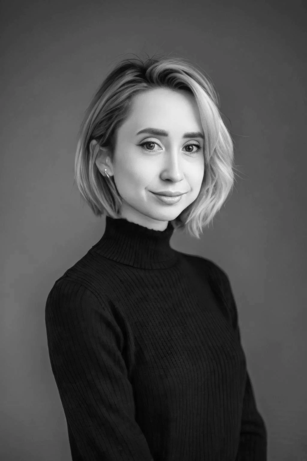 Porträt einer jungen Frau mit blondem Bob und schwarzem Rollkragenpulli, die ruhig und freundlich in die Kamera blickt – vor einem neutralen graugrünlichen Hintergrund.